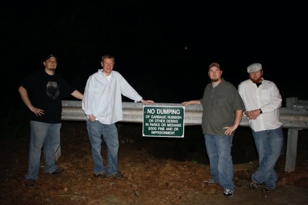 Four people standing beside a roadside sign at night.